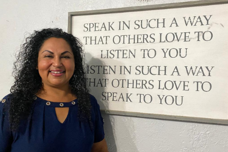 Nation of Neighbors Recipient Delfina Vazquez in front of a sign that reads, "Speak in such a way that others love to listen to you. Listen in such a way that others love to speak to you."